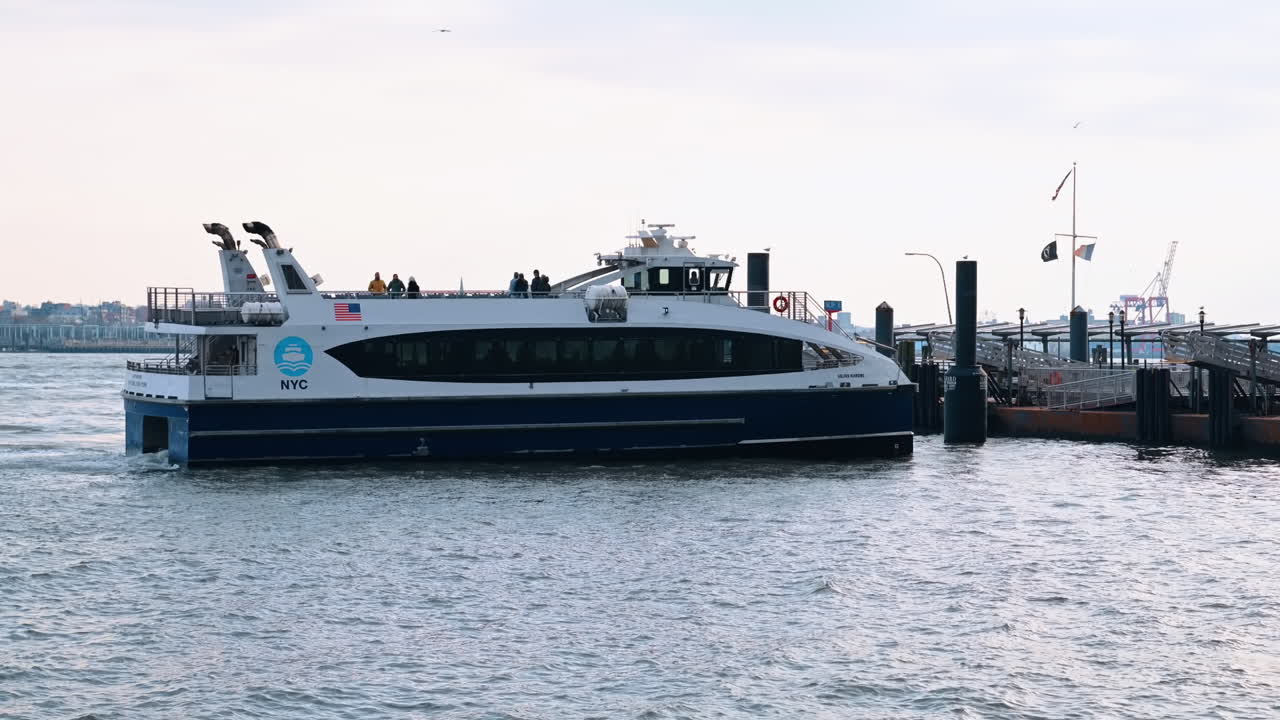 New York, USA, 5 dicembre 2025: traghetto NYC che si avvicina al molo sull'East River. Un traghetto NYC blu e bianco si dirige verso un molo sul fiume, con passeggeri visibili sul ponte e un vago skyline industriale