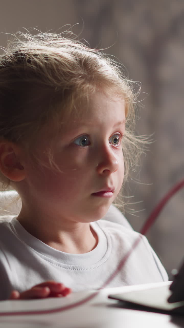 Emotional upset little girl with plait watches video lesson via tablet PC sitting at table in light children room close view slow motion