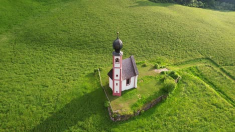 Aerial-tilt-down-view-of-San-Giovanni-Church-in-green-meadow-of-Funes-Valley,-Dolomites,-Italy