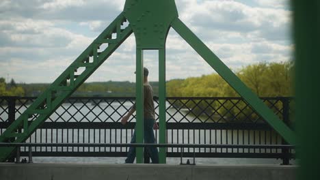 man walks over a truss bridge over a river and looks out into the distance with cars passing by