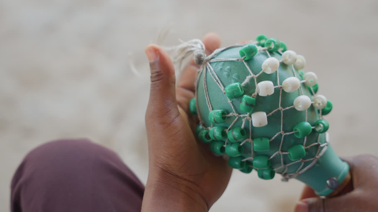 Nahaufnahme von schwarzen Händen, die einen Shaker halten. Grün-weißes Perlennetz um einen Kürbis-Shaker gewickelt, sandige Fingerspitzen und Handflächen, langsame sorgfältige Inspektion, texturierte Perlen und Knotenarbeiten in weichem natürlichem Licht.
