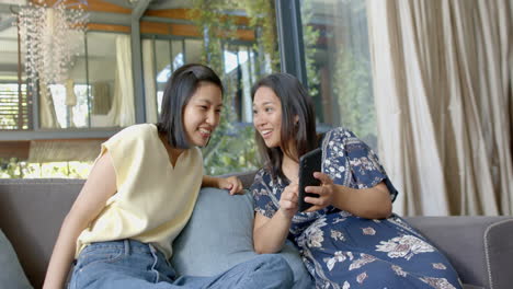 Happy-asian-female-friends-using-smartphone-and-smiling-in-sunny-living-room,-slow-motion