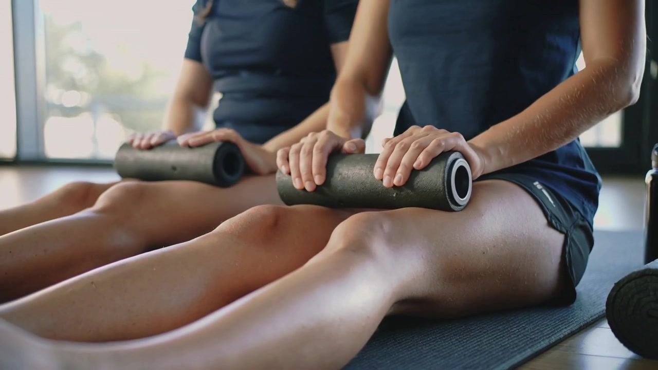 Começando com as pernas estendidas, duas mulheres de shorts escuros usando rolos de espuma pretos para recuperação em estúdio