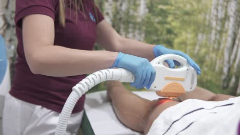 tracking shot of a professional cosmetologist using a hand-held laser hair remover on a patient's legs