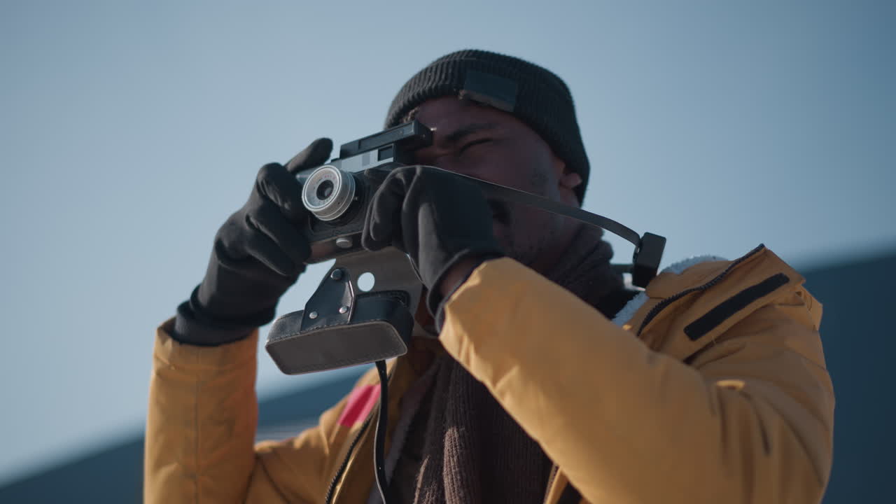 documentarian in yellow parka and knit beanie peering through camera viewfinder gloved hands fine tuning focus dial under clear blue urban winter sky bright sunshine over snowy plaza
