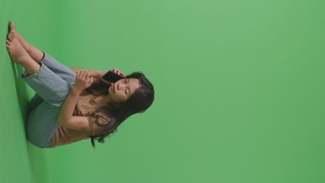 full body of young asian woman victim of violence with bruise on body sits in the green screen background studio crying and talking on smartphone asking for help