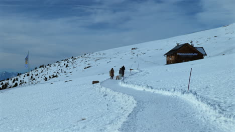 una familia camina por un camino nevado en las montañas cerca de una bandera bávara y una cabaña de madera