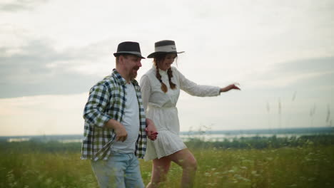 a cheerful couple holds hands and runs through a grassy field at sunset. the man, wearing a black hat and checkered shirt, and the woman, in a white dress and matching hat