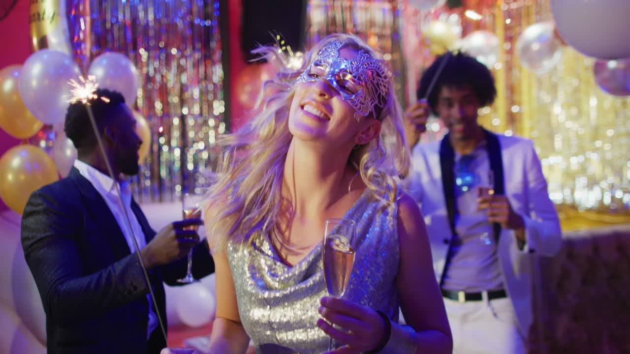 Video of happy caucasian woman in mask dancing with sparkler and glass of champagne at a nightclub