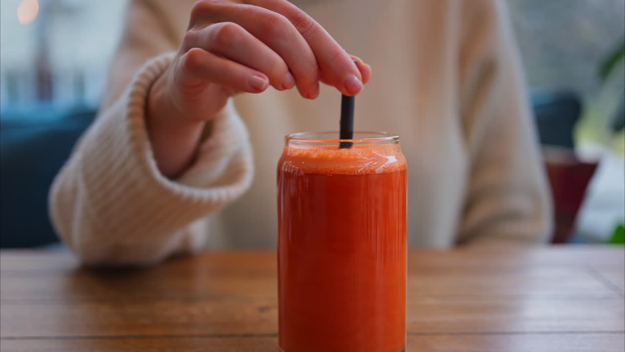 Close de uma mulher misturando suco de laranja e cenoura em um copo com um canudo preto sobre uma mesa de madeira