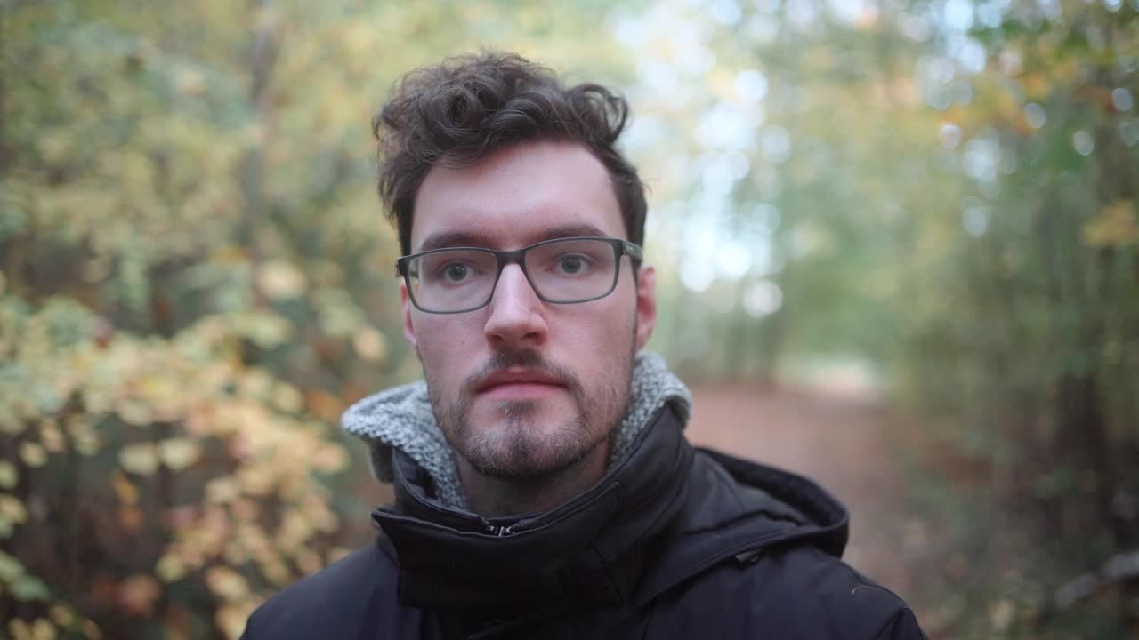 en cautivadora cámara lenta, un joven europeo con gafas afiladas y una barba elegante encuentra consuelo en un bosque mixto de otoño, su mirada atravesando el lejano horizonte, perdido en sus pensamientos