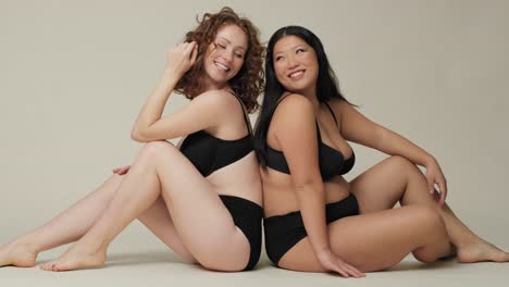 two women in underwear sitting back to back on the floor in the studio.