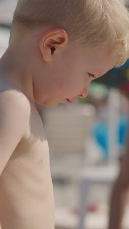 curious blond toddler boy with bare torso walks along blurry crowded public beach at seaside resort on sunny summer day close view slow motion