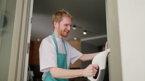 Side-view-of-a-confident-blond-guy-in-a-white-T-shirt-and-blue-apron-washes-a-glass-door-using-a-window-vacuum-cleaner-during-a-gentle-cleaning-of-the-apartment