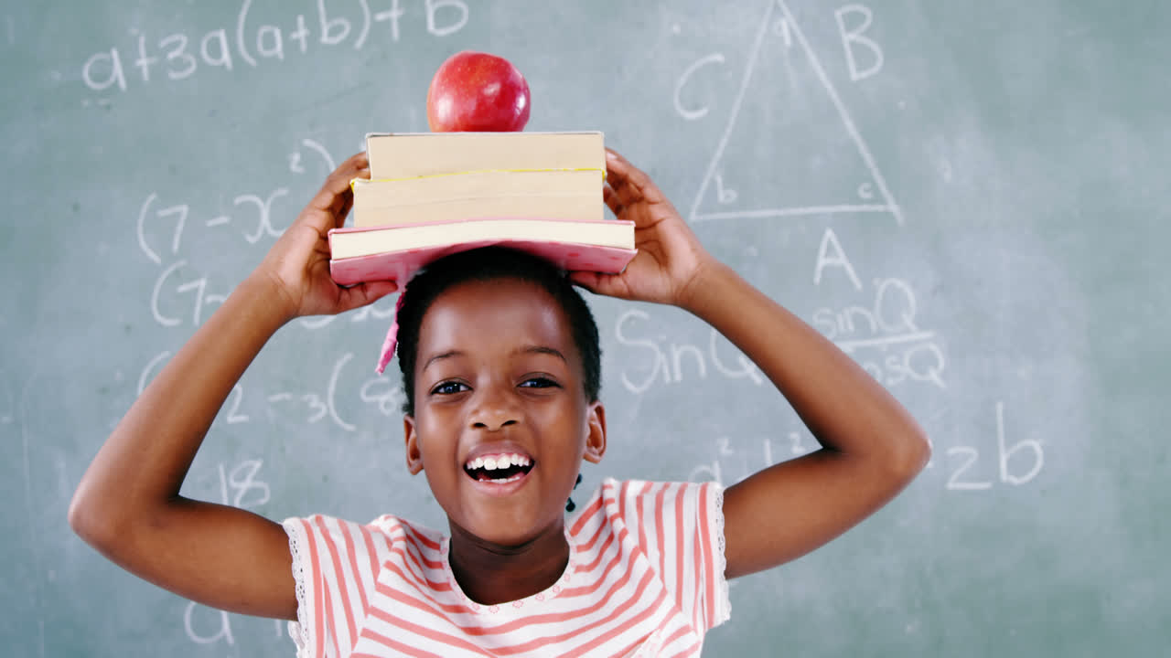 Schoolgirl holding books stack with apple on head against chalkboard