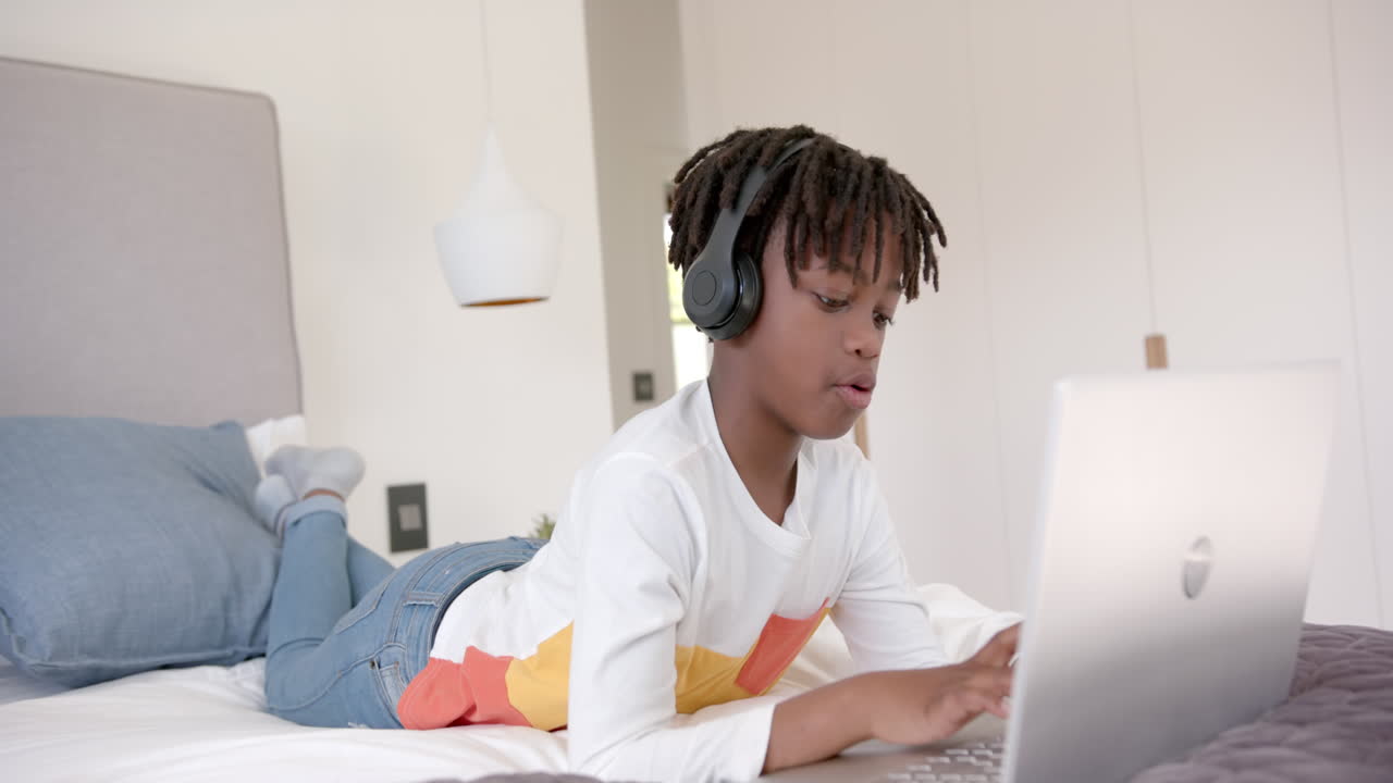 Happy african american boy wearing headphones lying on bed using laptop at home, slow motion