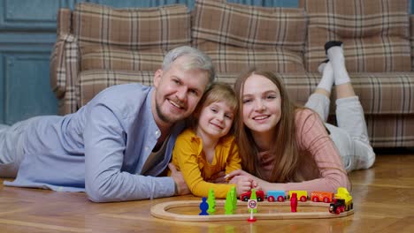 Familie-Aus-Mutter,-Vater-Und-Tochter,-Die-Im-Wohnzimmer-Auf-Einer-Holzeisenbahn-Mit-Einer-Spielzeugeisenbahn-Fährt