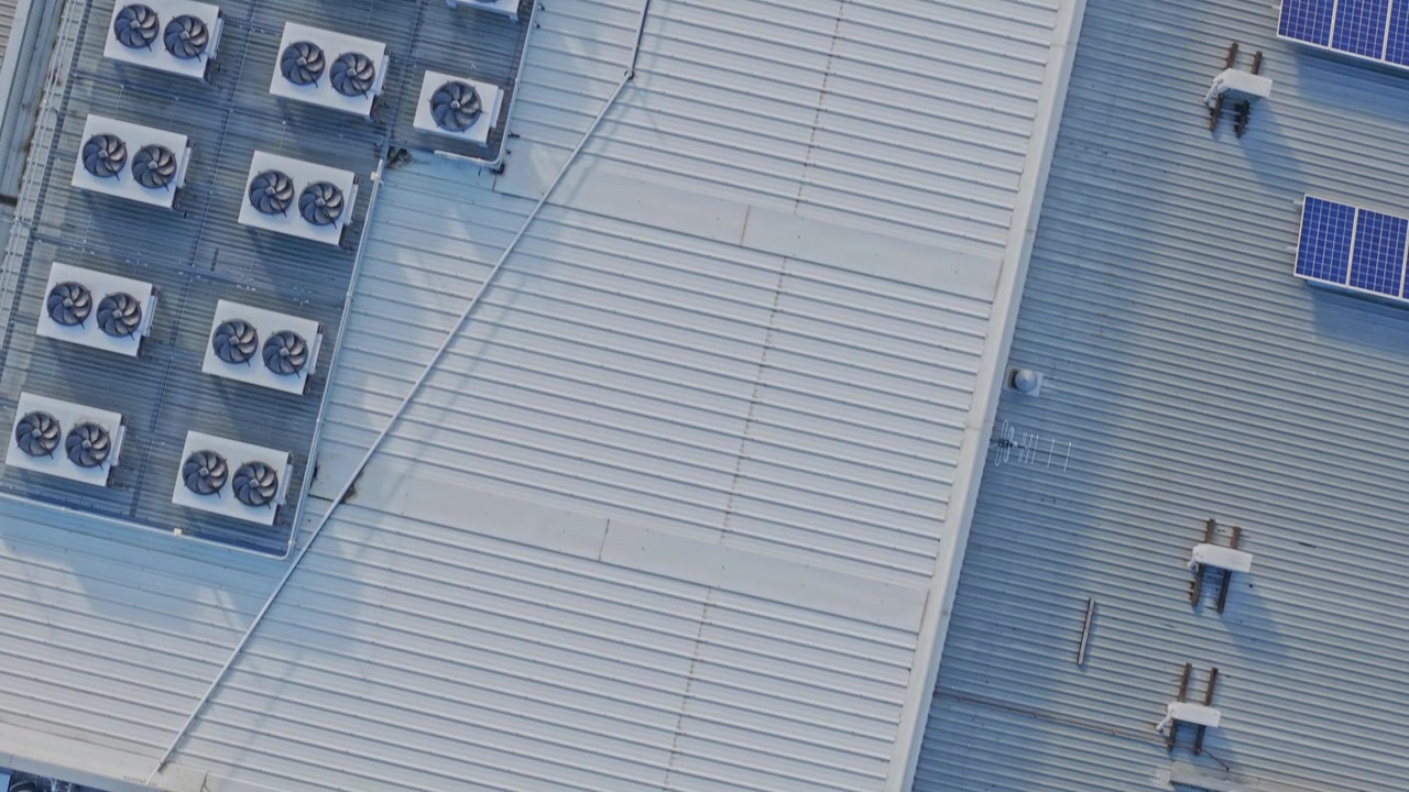 Drone footage shows a sweeping overhead shot of a high-tech data centre roof, featuring neatly arranged HVAC units and blue solar panels on a metallic surface in bright daylight.