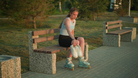 joven sentada al aire libre en un banco con zapatillas blancas en la mano derecha mientras cae en el suelo mientras usa patines, con árboles y vegetación en el fondo