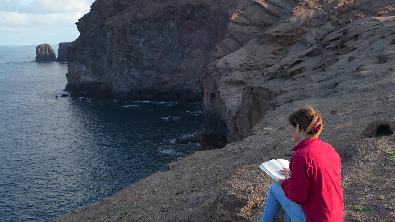 Woman reading book while sitting on rocky cliff, embracing serene ocean landscape of Gran canaria with peaceful contemplation and vibrant natural surroundings