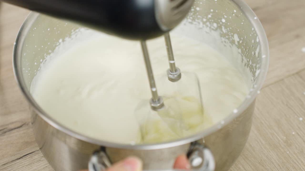 Person using hand mixer on homemade whip heavy cream in a stainless steel bowl in kitchen with light wood surface top