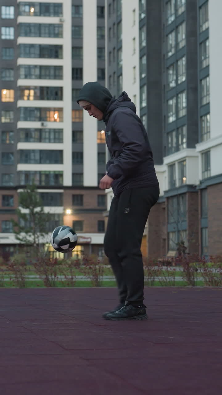 Coach skillfully juggling football on sport arena surrounded by urban scenery and modern architecture, background features people walking near buildings, greenery, and evening lights