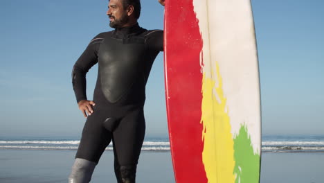 vertical shot of a male surfer in wetsuit with artificial leg standing on the beach while leaning on the surfboard and looking in front of him