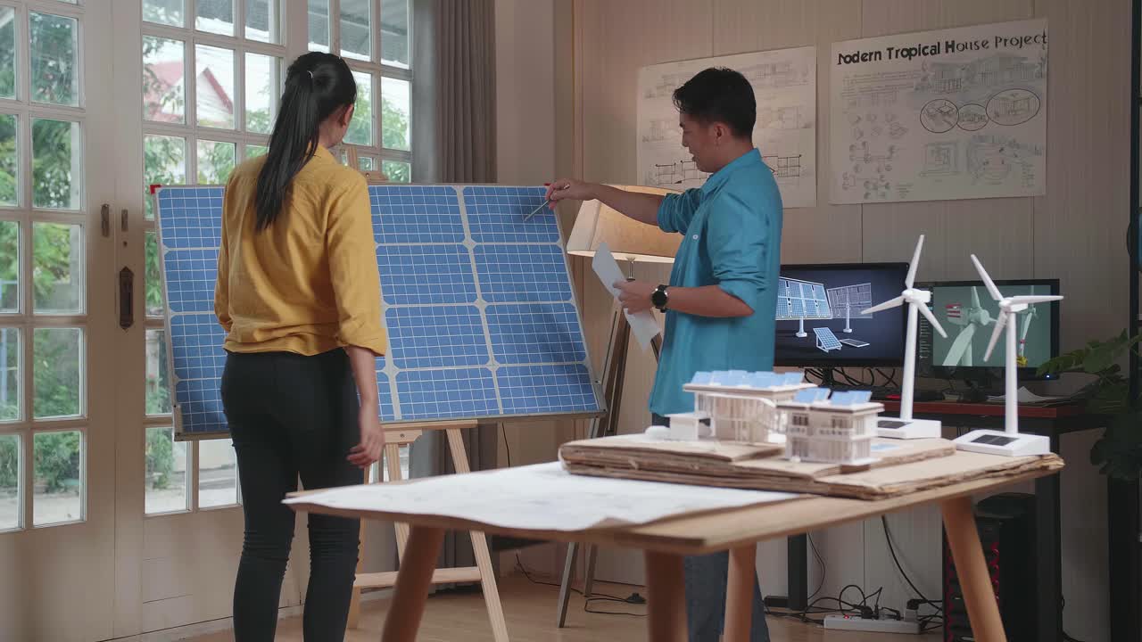 아시아 남녀가 태양전지에 대해 논의하는 방으로 걸어 들어가며 태양 패널 모형 집 에 청사진이 있고 테이블 위에 풍력 터빈이 있습니다.