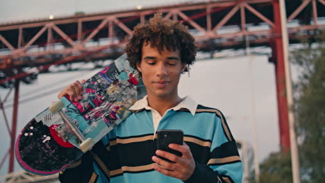 contemporary skateboarder texting phone closeup. curly man carrying longboard