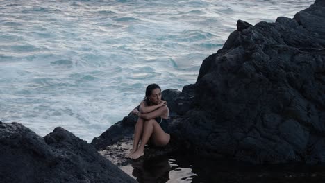 a woman sits quietly on volcanic rocks beside the atlantic ocean in seixal, madeira, surrounded by waves and reflective stillness.