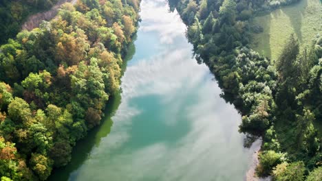 Camino-Rural-A-Lo-Largo-De-Un-Río-Azul-Brillante-Bajo-Un-Sol-Brillante-Rodeado-De-Bosque,-Drones-Aéreos