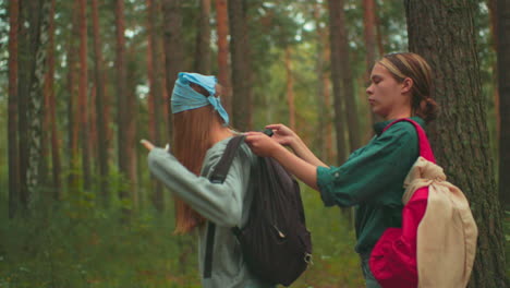 young woman in red backpack assists her friend wearing a black bag in peaceful forest, friend in blue scarf looks back with a smile as they prepare to walk, surrounded by serene trees and greenery