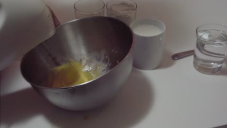 Mixing-eggs-with-sugar-in-stainless-steel-bowl-for-pancake-batter---ingredients-on-table-for-recipe