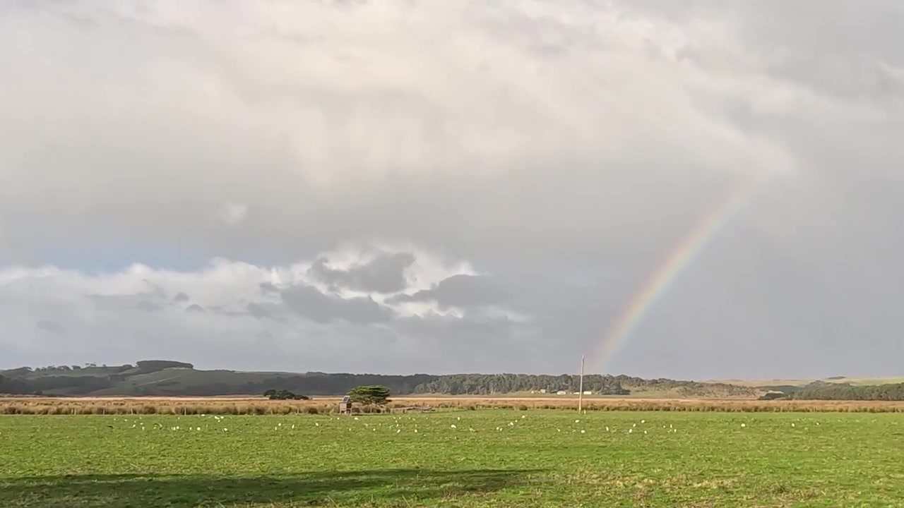 En färgstark regnbåge sträcker sig över en molnig himmel över vidsträckta gröna fält och landsbygd.