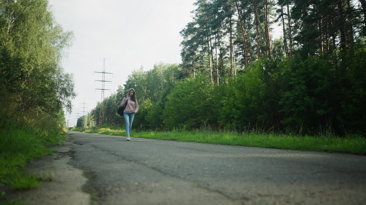 Young lady in pink hoodie walking along quiet forest road with one hand in pocket and black bag on shoulder, surrounded by lush greenery, trees, and distant power poles under soft daylight