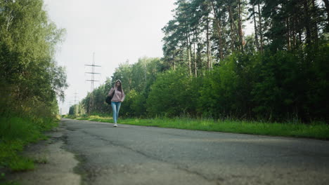young lady in pink hoodie walking along quiet forest road with one hand in pocket and black bag on shoulder, surrounded by lush greenery, trees, and distant power poles under soft daylight
