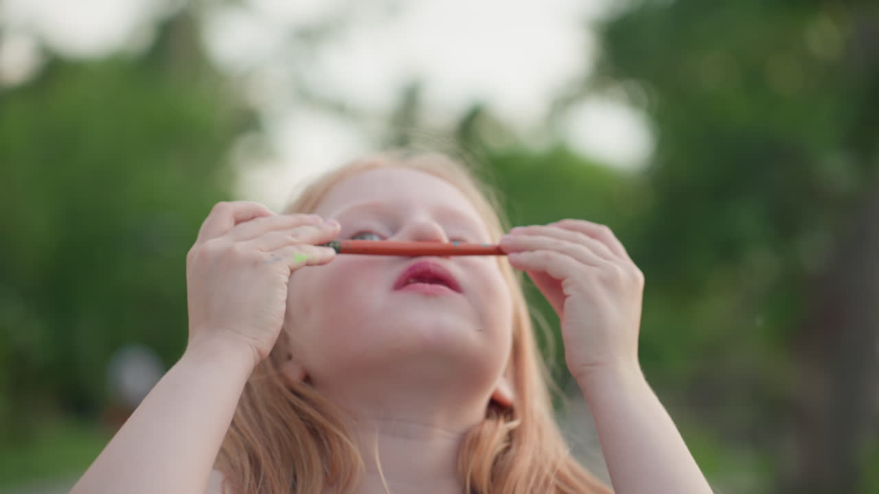 close up young girl holding paint brush under nose sniffing colorful paint playfully with eyes closed and lips puckered while seated outdoors by water against blurred green trees in warm evening