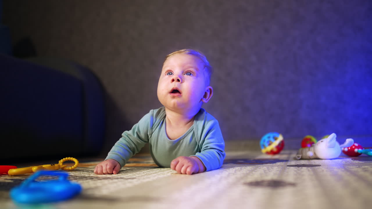 Adorable Caucasian baby boy with big blue eyes lies on the floor on his belly. Lovely infant looking up with surprise and smiling.