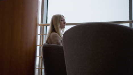 reflective lady in woven sweater, seated on modern chair in mall, looking contemplative. iron rail and soft lighting in background, creating a peaceful, introspective atmosphere in a stylish setting