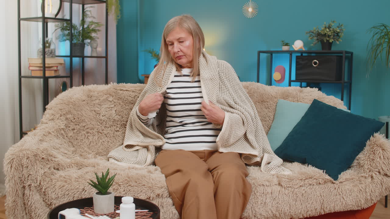 Mature woman covered with blanket on sofa checking temperature with thermometer wipes snot at home
