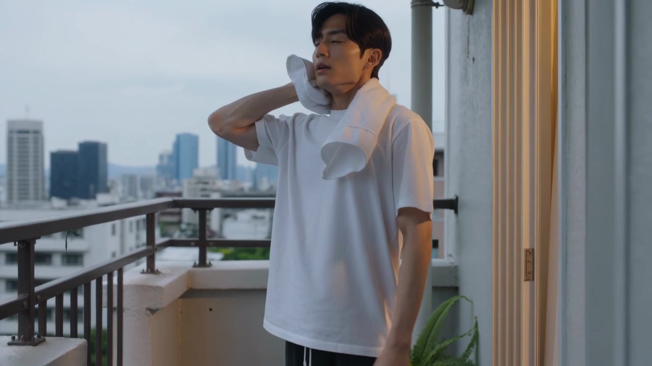 Uomo asiatico in maglietta bianca che esce da una porta, asciugandosi il viso con un asciugamano su un balcone, mentre si rinfresca.