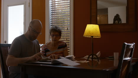 a middle-aged man, and elderly woman sit together at a dining table in a warmly lit home, sorting through receipts. son is focused on counting and calculating receipts, onlooking mother disapproves.