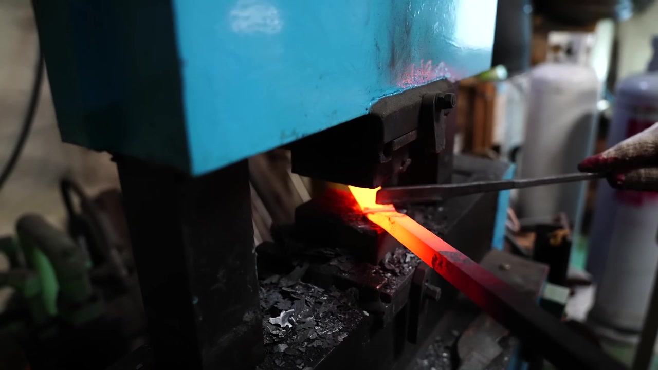 Close up of a glowing steel bar tapered under a forging press die inside a Korean knife making workshop
