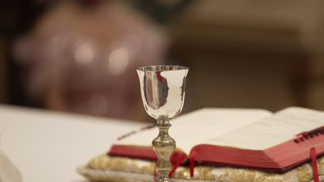 priest's hand turning page of prayer book next to silver chalice during mass