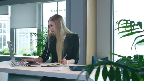 elegant woman working in stylish office. modern blond woman in trendy suit sitting at table in light contemporary office and writing on papers