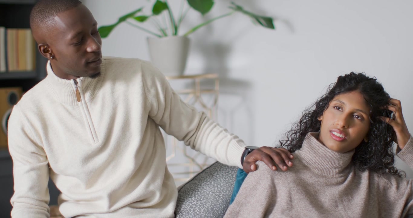 Diverse couple on sofa, man turning leaning placing hand on her shoulder comforting football on lap