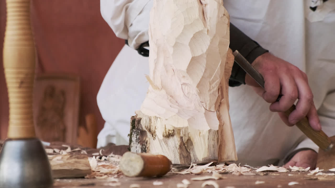 Static close-up of an artisan carving a log with a chisel; foreground tools and chips as shavings fly and fall across the workbench, warm daylight, shallow depth of field