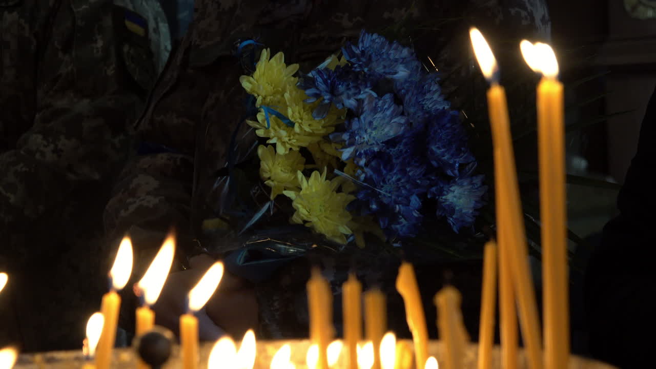 un soldado que sostiene flores azules amarillas y azules se ve a través de velas de oración encendidas en el funeral de un soldado ucraniano durante la invasión rusa del país
