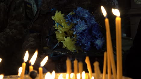 un soldado que sostiene flores azules amarillas y azules se ve a través de velas de oración encendidas en el funeral de un soldado ucraniano durante la invasión rusa del país