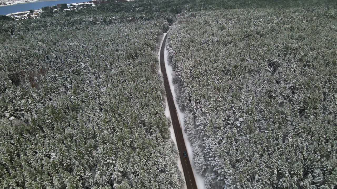 drone shot of the narrow asphalt road surrounded by snow-covered forest pine trees on a cloudy frosty winter day